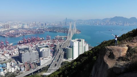 Woman stay on rock at top, stretch up hands, camera fly over. Hong Kong city panorama and large Stonecutters bridge seen ahead below. Hiker girl enjoy beautiful scenery from viewpoint at mountain