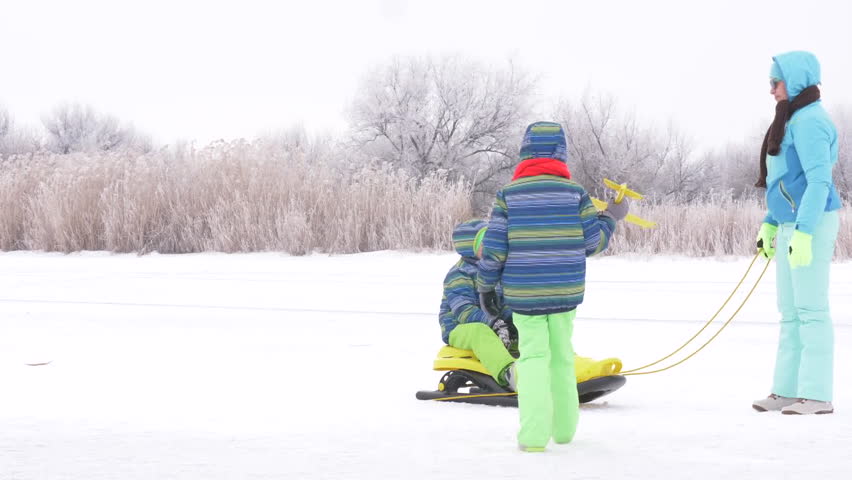 Mom in blue ski suit and three happy children in warm winter clothes ride on yellow sled on frozen snow-covered river.  Concept of friendly sports family. Winter holidays in village. Christmas eve