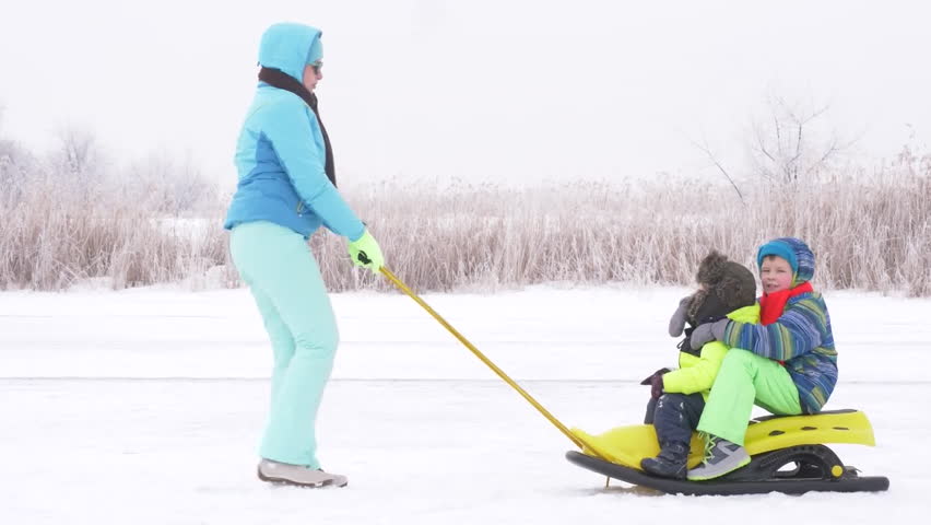Mom in blue ski suit and three happy children in warm winter clothes ride on yellow sled on frozen snow-covered river.  Concept of friendly sports family. Winter holidays in village. Christmas eve