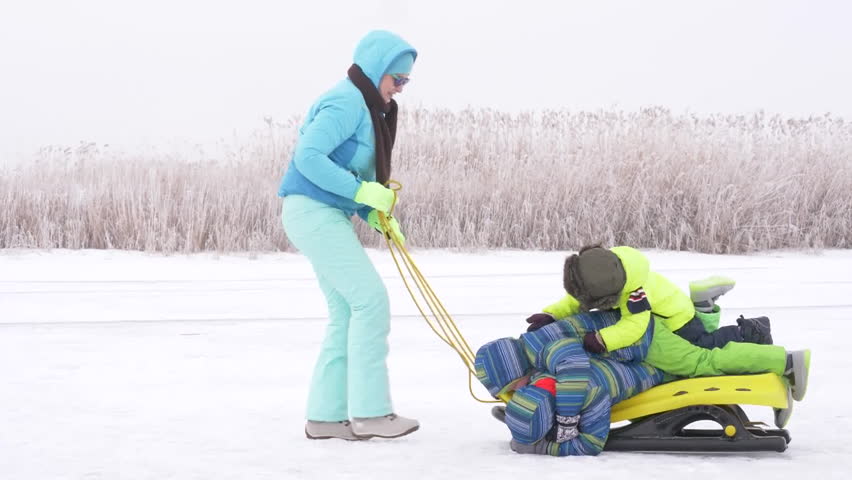 Mom in blue ski suit and three happy children in warm winter clothes ride on yellow sled on frozen snow-covered river.  Concept of friendly sports family. Winter holidays in village. Christmas eve