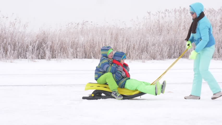 Mom in blue ski suit and two happy children in warm winter clothes ride on yellow sled on frozen snow-covered river.  Concept of friendly sports family. Winter holidays in village. Christmas eve