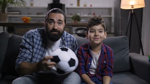 Overjoyed father with mixed race son rejoicing at goal with fists clenched, happily shouting while watching soccer game on tv. Excited football fans celebrating victory of soccer team at home.