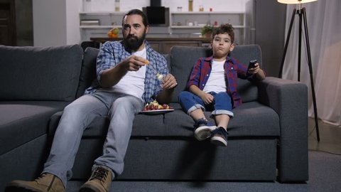 Relaxed bearded single father and cute mixed race son with remote control watching movie on tv and eating delicious fruit kebabs while enjoying freetime together on sofa in domestic room.