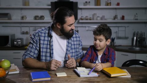 Positive loving stay-at-home bearde father writing on sticky note paper household chores while giving housework tasks to his mixed race elementary age son in domestic kitchen.