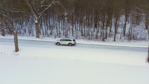 Tracking shot of an estate car travelling on a snowy country road. Side tracking shot of white car during winter with trees and small house in the background. Passing over a stream into a forest.