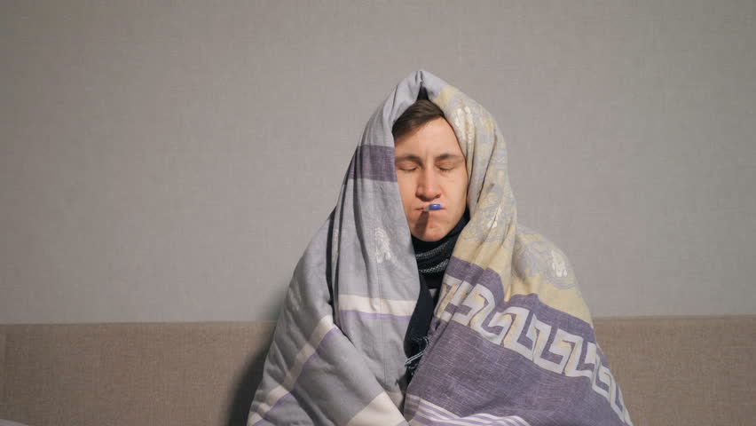 Unhappy young man wrapping in warm blanket and keeping electronic thermometer in mouth while suffering from illness at home