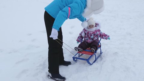 Young mother rolls little baby on sled along snowy road in winter. Child is naughty and cries while sitting in sled.