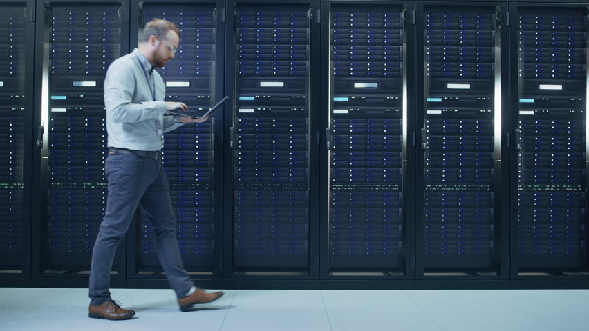 Ginger Bearded Data Center IT Technician in Glasses is Walking Through Server Rack Corridor with a Laptop Computer. He is Visually Inspecting Working Server Cabinets. | Shutterstock HD Video #1021974541