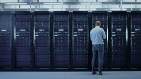 Bearded IT Specialist in Glasses is Working on Laptop in Data Center while Standing Before Server Rack. Running Diagnostics or Doing Maintenance Work. Camera Zooms Out. - Powered by Shutterstock - Get 15% off with code: PIKWIZARD15