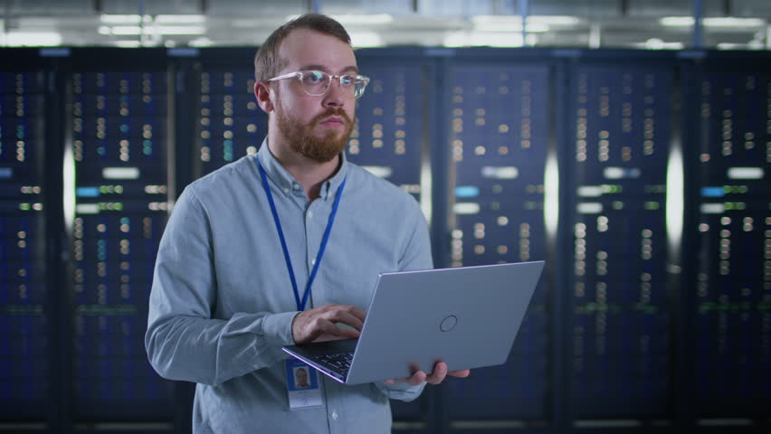 Bearded IT Specialist in Glasses is Working on Laptop in Data Center Next to Server Racks. Running Diagnostics or Doing Maintenance Work. | Shutterstock HD Video #1021974604