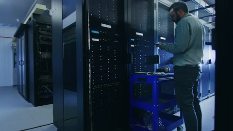 Black IT Technician with a Laptop Computer Gives a Tour to a Young Intern. They Talk in Data Center while Walking Next to Server Racks. Other Colleague is Changing Hard Drives.