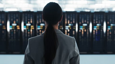 Follow Back Shot of a Female IT Chief Engineer in a Jacket Walking Towards a Server Rack in Data Center Room.