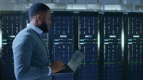Black Data Center IT Technician Walking Through Server Rack Corridor with a Laptop Computer. He is Visually Inspecting Working Server Cabinets. - Powered by Shutterstock - Get 15% off with code: PIKWIZARD15