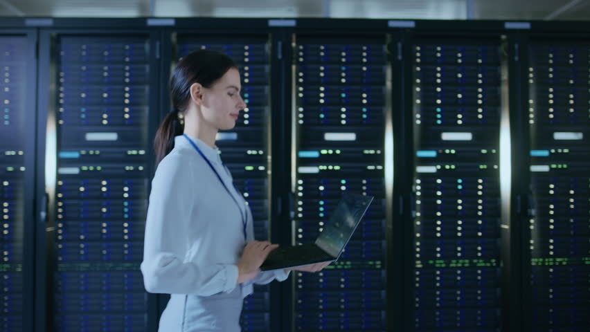 Beautiful Data Center Female IT Technician Walking Through Server Rack Corridor with a Laptop Computer. She is Visually Inspecting Working Server Cabinets. | Shutterstock HD Video #1021974820