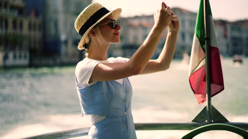 Positive hipster girl in stylish hat enjoying time for recreating during italian weekend taking video of architecture using camera on modern mobile phone while floating on touristic boat with flag