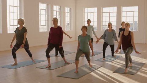 yoga class of healthy mature women practicing warrior pose enjoying morning physical fitness exercise workout in studio at sunrise