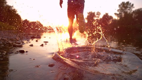 SUPER SLOW MOTION, CLOSE UP Unrecognizable young man running in river shallows at golden sunset. Active sportsman having a morning run in shallow water. Male runner jogging, splashing water at sunrise