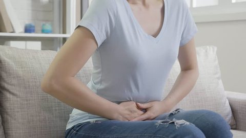 Stomach pain or menstrual pain. unrecognized asian woman with pains in abdomen sitting on couch wearing t shirt in living room at home. Female hands holding touch belly moving front and back.