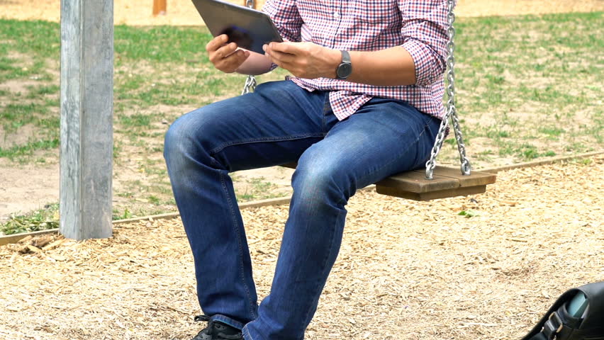 Young man watching movie on tablet sitting on swing in city park