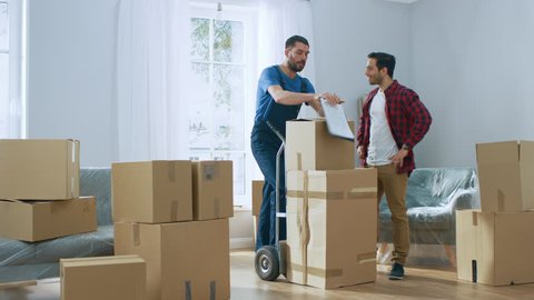 Happy New Homeowner Welcomes Professional Mover with Hand Truck full of Cardboard Boxes, Receives His Goods and Signs on Clipboard.