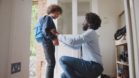 Middle aged black man wearing a shirt and tie kneeling down preparing his son and saying goodbye before he leaves home for school in the morning, side view