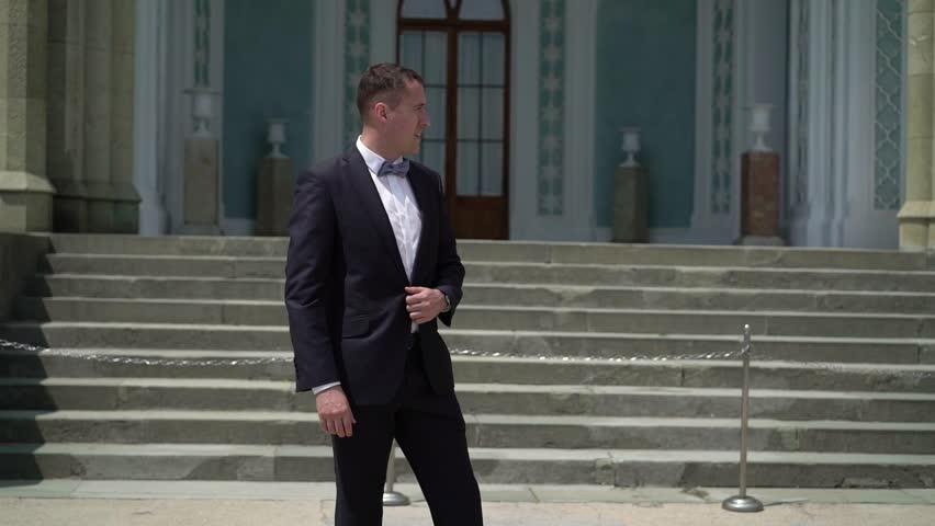 Young man in suit waiting, staying and posing in a city at windy summer day. Slowmotion