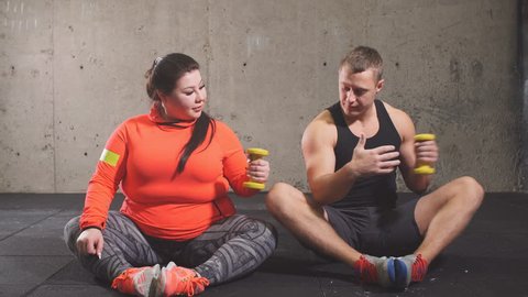 Positive overweight woman and fit guy doing exercises while sitting on the floor. Funny learning.