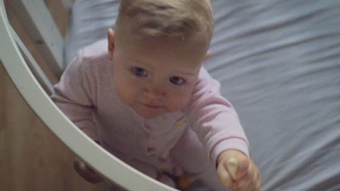 A top view of a smiling baby girl in pink clothes, sitting in a crib and looking up with big beautiful eyes. She is holding the railing and trying to stand up
