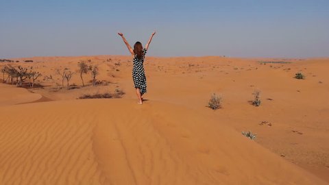 AERIAL. Woman standing on top of dune in desert and looking at rising sun in mist with hands up, travel in UAE Dubai