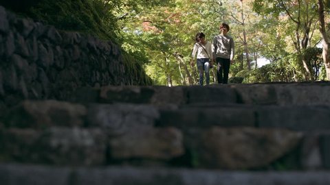 Young couple walking down a stone pathway towards steps under a shaded forest in Kyoto, Japan with soft natural lighting. Medium shot on 4k 