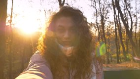 Self-video of caucasian curly-haired woman in sunshine making selfie-photos in autumnal park.