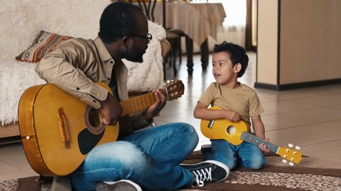 African American man sitting on carpet at home near his biracial son, both playing guitars and singing song