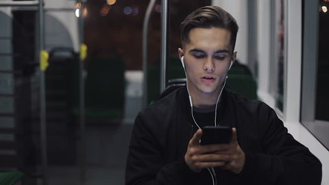 Portrait of young headsome men in headphones listening to music and browsing on mobile phone in public transport. City lights background. Slow motion