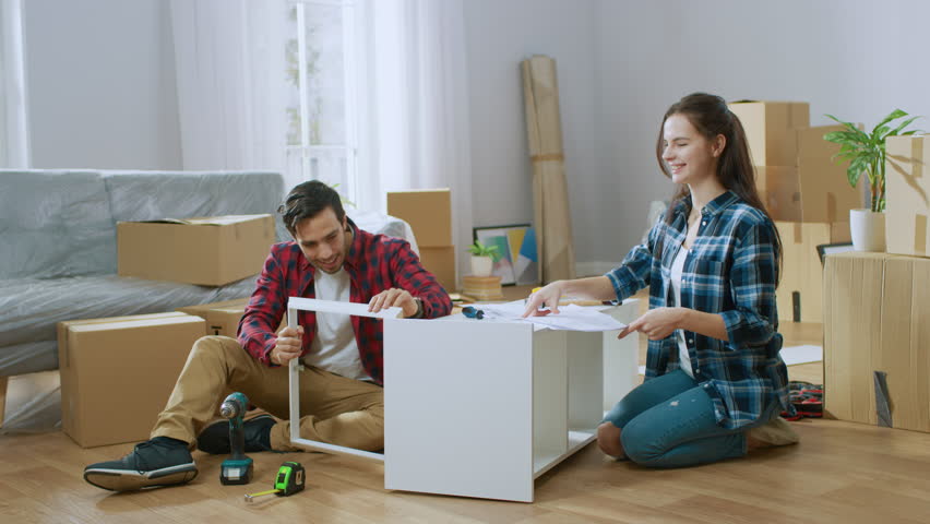 Happy Couple Assembles Furniture as a Team, Girl Reads Instructions and Boy Tightens Screw with a Drill. Moving into New Apartment, Couple Assemble Shelf. Royalty-Free Stock Footage #1022087905