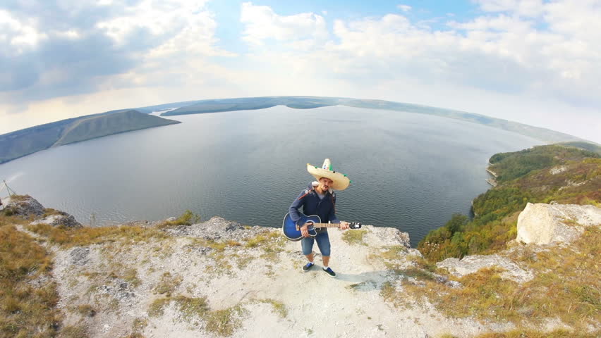 Handsome mexican mariachi playing acoustic guitar and singing on the green hills over the large lake in summer. Fisheye, camera movement, inspiring sight.
