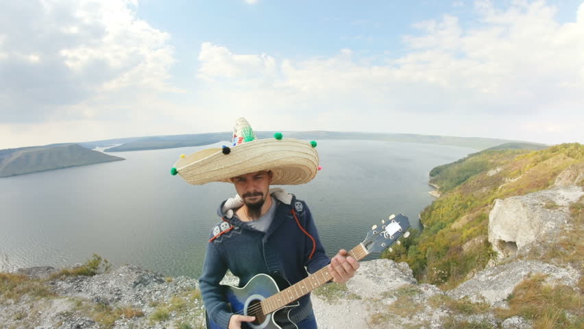 Fisheye view of Mexican boy shaking his sombrero while singing and dancing over the stunning lake and hills.