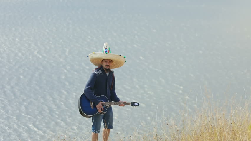 Attractive young man wearing sombrero singing and playing guitar over the huge peaceful lake.