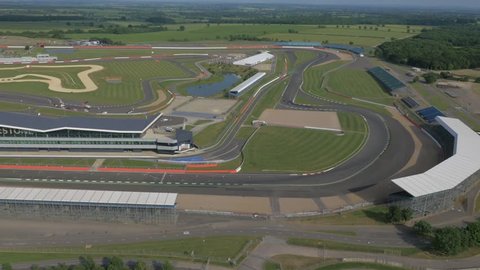 Winding racetrack with grandstands and pit area. Two cars make their way round the circuit at speed, wide aerial panning perspective