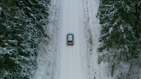 Estate car driving through a narrow country road in a dense spruce forest aerial. White car driving in a dense forest turning right onto a larger snowy country road