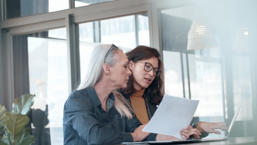 Two business woman working together in office with reports and laptop. Senior executive with colleague looking at business financial report.
