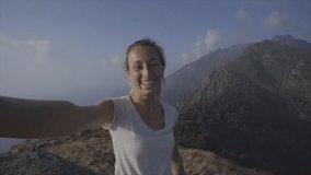 Cheerful young woman taking a selfie portrait on mountain top after hiking all the way up reaching summit; People enjoying Summer outdoor activities concept 