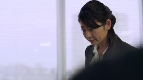 Medium shot on 4k RED camera. Successful Japanese woman observing a group of coworkers sitting and talking in an office with soft interior lighting.