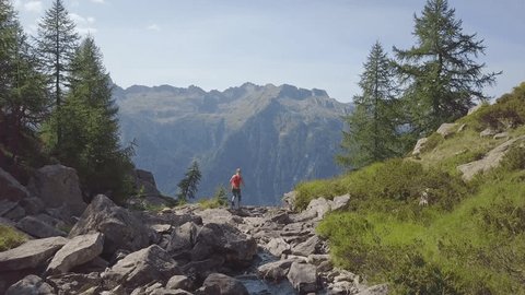 Woman walking following river, stops and looks at view from above stunning waterfall. Drone shot, aerial view of woman hiking in the mountains 
