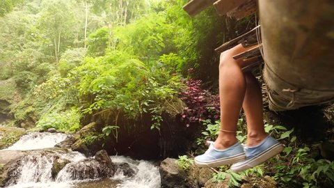 Young Traveler Girl Relaxing by the Jungle Waterfall, Sitting on Wooden Bamboo Bridge and Dangle with Her Feet. 4K Slowmotion. Bali, Indonesia.