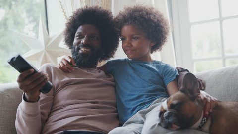 Smiling middle aged black father and pre teen son reclining on sofa together watching TV with their pet dog, dad holding remote control, low angle, close up