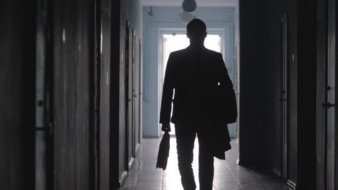 Tilt down shot of silhouette of businessman wearing formal suit and carrying coat and briefcase leaving office and walking towards the camera along hallway 