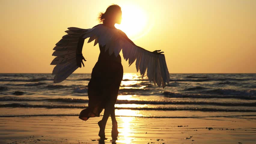 Silhouette of young girl with angel wings dancing at sunset. Silhouette of female artist is dancing on the sandy beach against orange sunset, slow motion