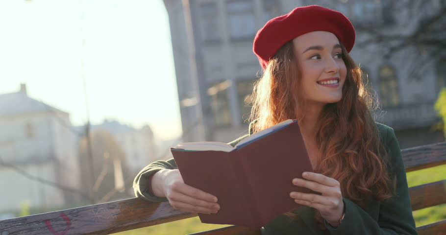 Portrait of the Caucasian beautiful youg girl with red hair and in a cap resting on a bench outdoors with a book, then looking at the side and dreaming.