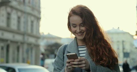 Young Caucasian red-hair student girl chatting and texting on the phone device while smiling and stepping the city street.
