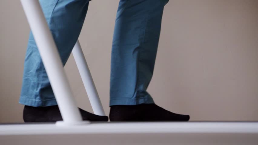 View of metallic medical device track with person's legs wearing blue pants and black socks slowly walking on it in room with white walls.